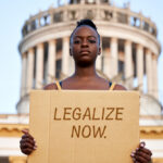 A woman in front of the Capitol building holds up a sign demanding cannabis legalization.