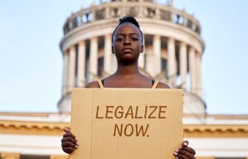 A woman in front of the Capitol building holds up a sign demanding cannabis legalization.